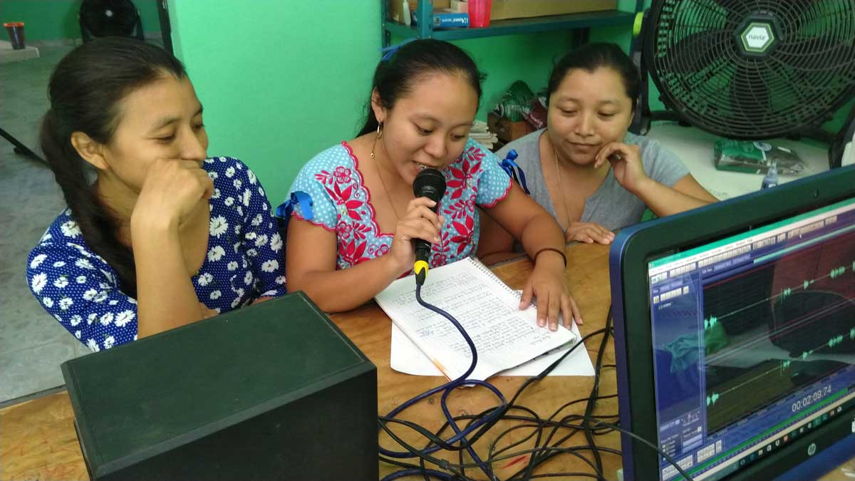 Three women collaborating on an audio recording with a computer and microphone.