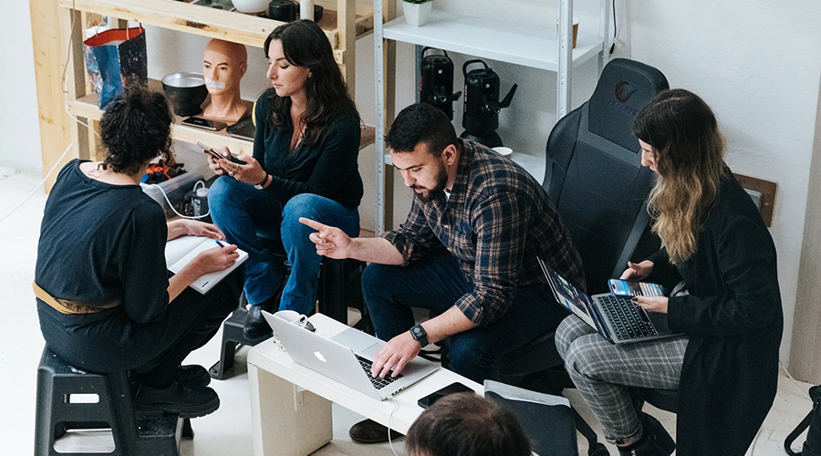 A group of people collaborating in an informal workspace with laptops and phones, surrounded by shelves with various items.