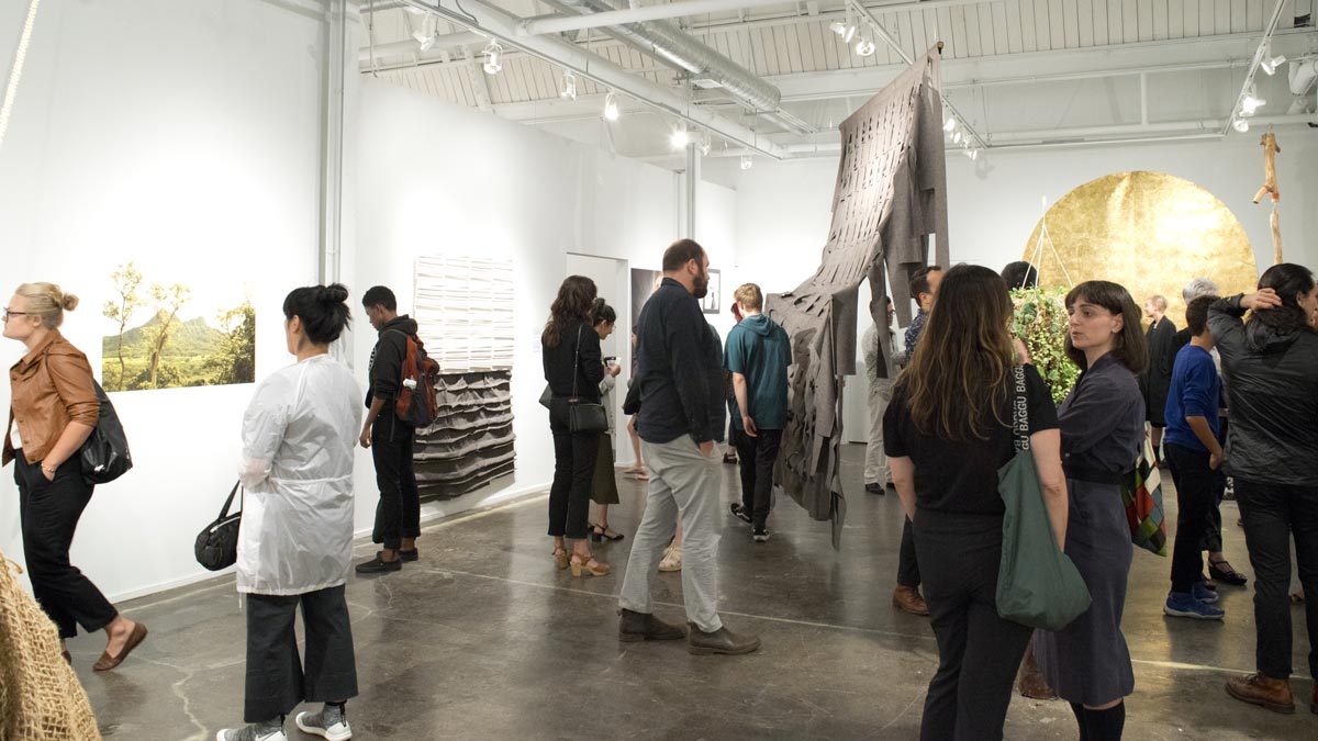 People view artwork in a contemporary gallery with white walls, showcasing photographs, textured pieces, and a large fabric sculpture.