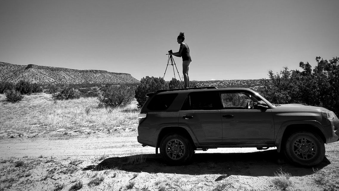 a black and white photo of a woman with a camera tripod, standing on top of a truck parked in the desert.