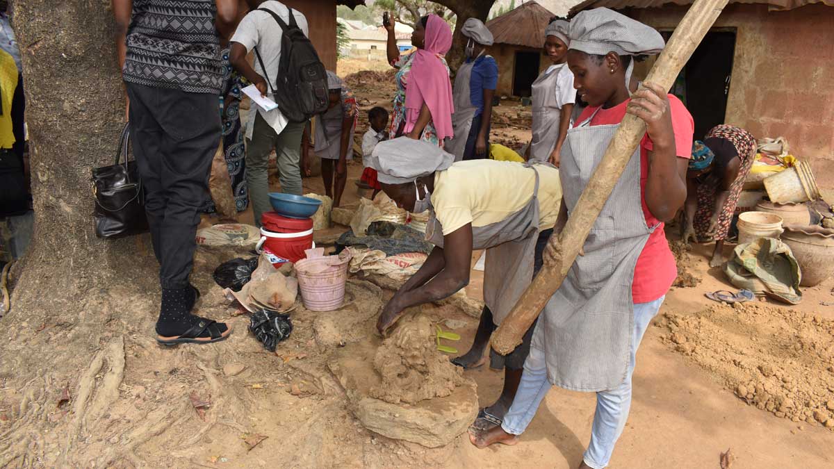 People in aprons make pottery under a tree, with clay and tools, in an outdoor setting. Some others are gathered nearby.