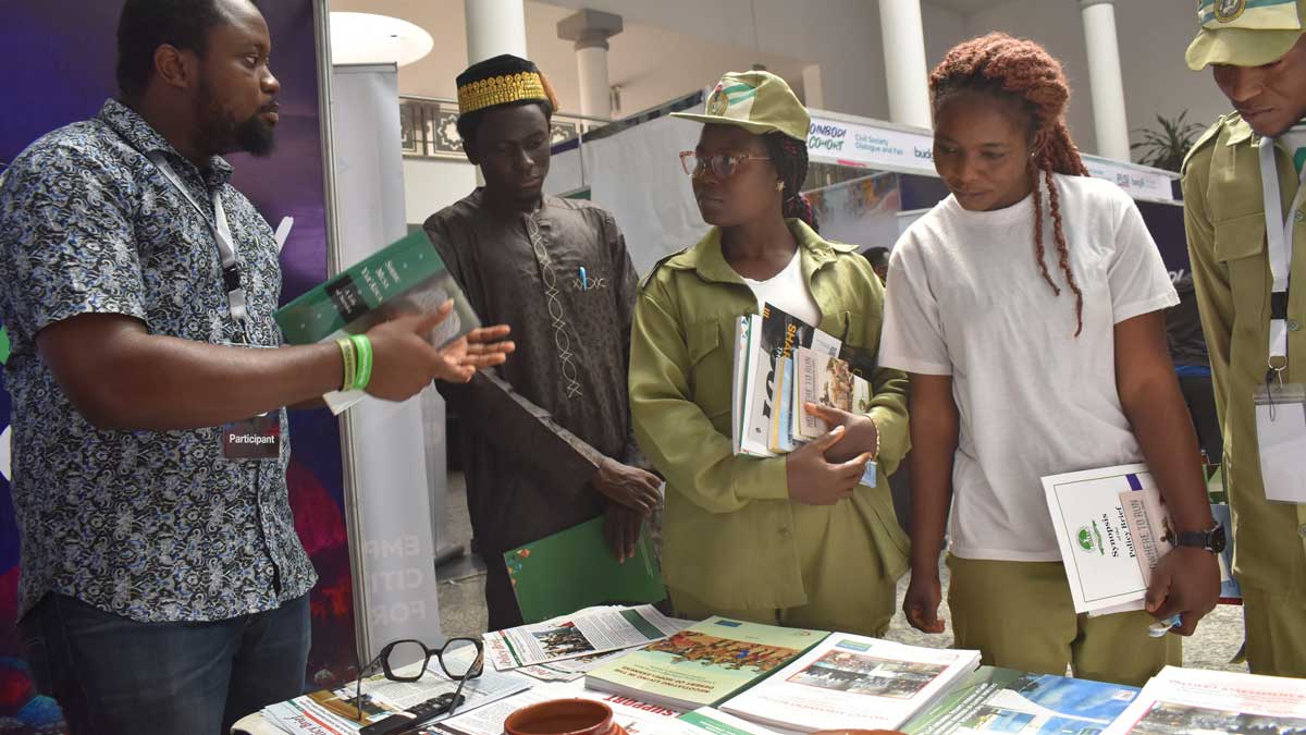 A group of young people at a booth, engaging with a presenter at an event, holding various informational materials.