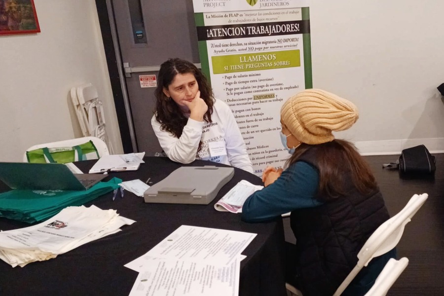 A woman with brown hair sits at a table listening to another woman in a beige hat sitting across from her. 