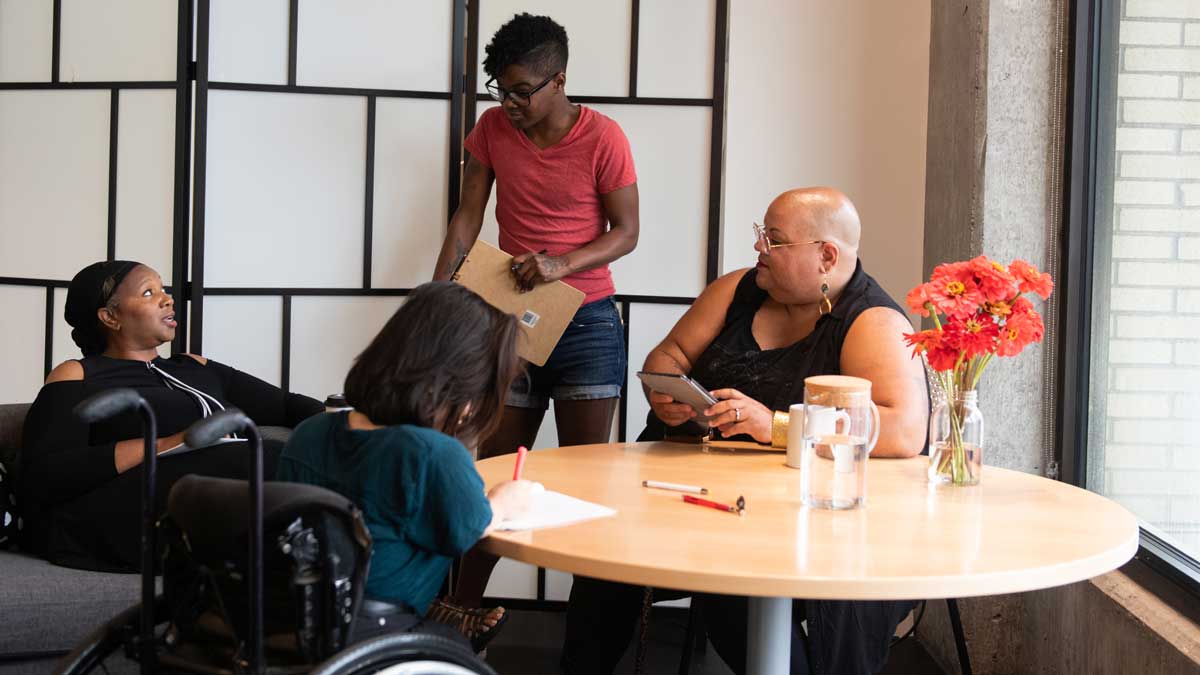 Four disabled people of color gather around a table during a meeting. A Black woman sitting on a couch speaks with a serious expression while the three others (a South Asian person sitting in a wheelchair and taking notes, a Black non-binary person sitting in a chair with a tablet and cane, and a Black non-binary person standing with a clipboard) listen.