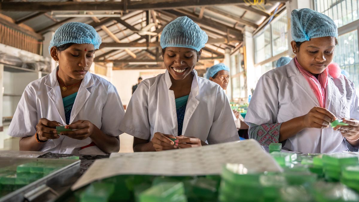 Three women in lab coats and hairnets work on packaging items in a bright, industrial setting.