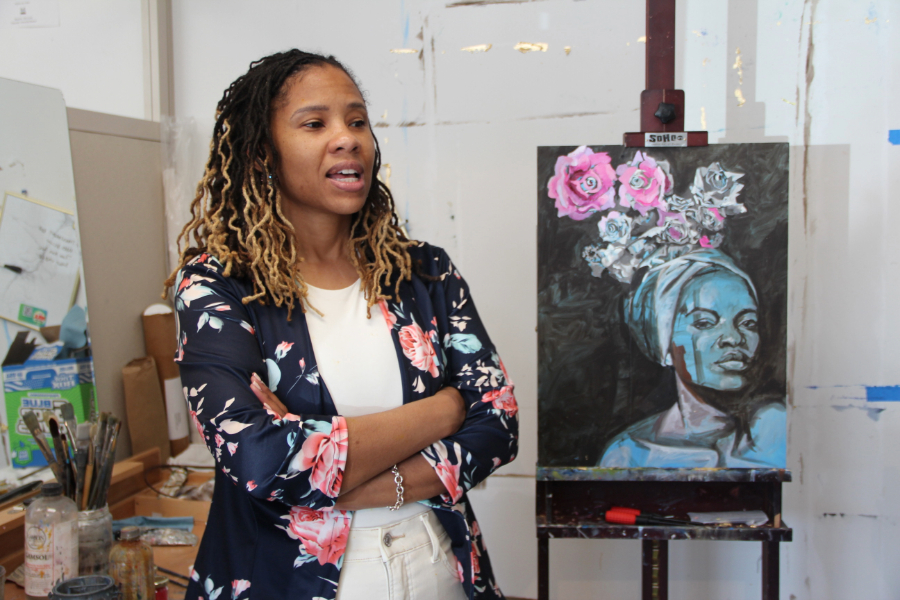 An artist stands and speaks in front of a painting of a woman in an art studio.