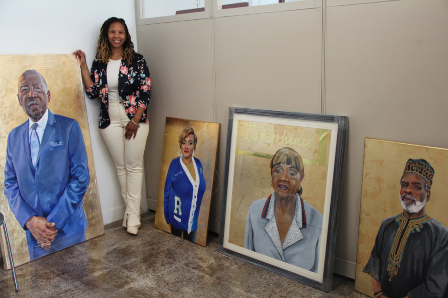 An artist stands next to portraits they have painted. 