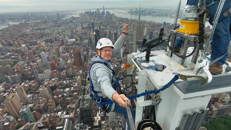 A man wearing a helmet and harness hanging off the side of a building takes a selfie.